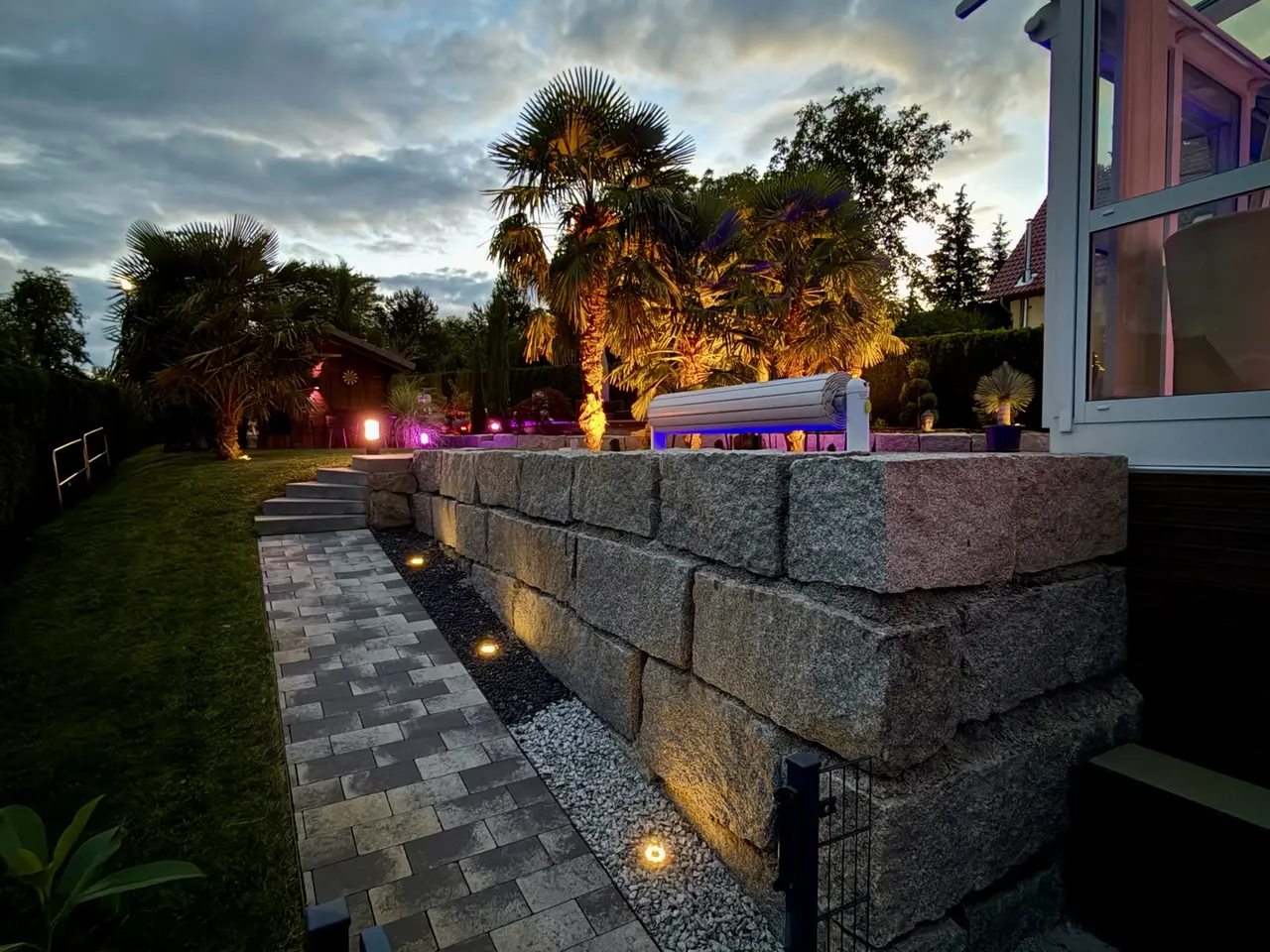 Beleuchtete Natursteinmauer mit Gartenweg und Palmen am Abend