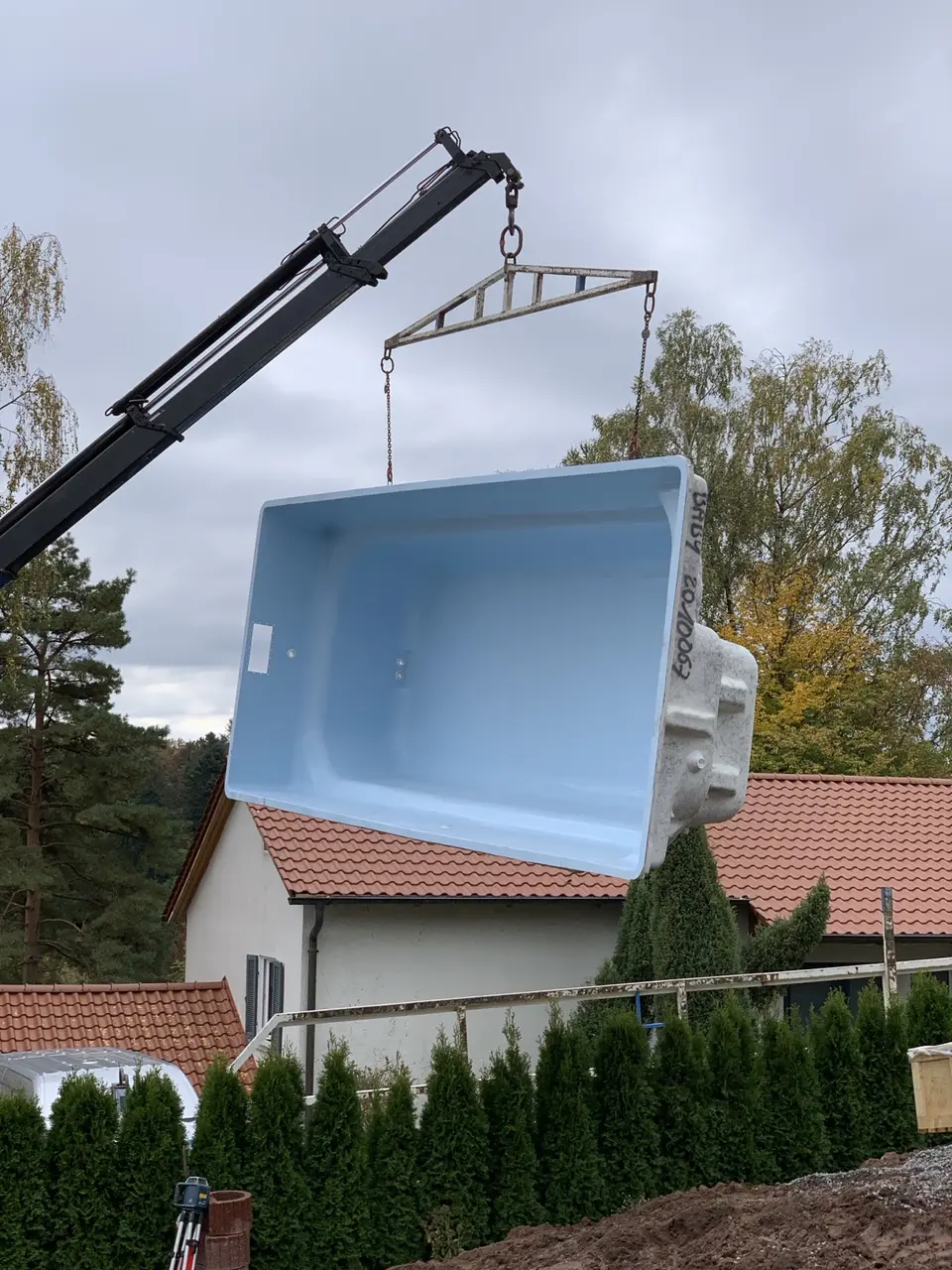 Ein hellblaues Fertigbecken wird mit einem Kran über die Hecke gehoben