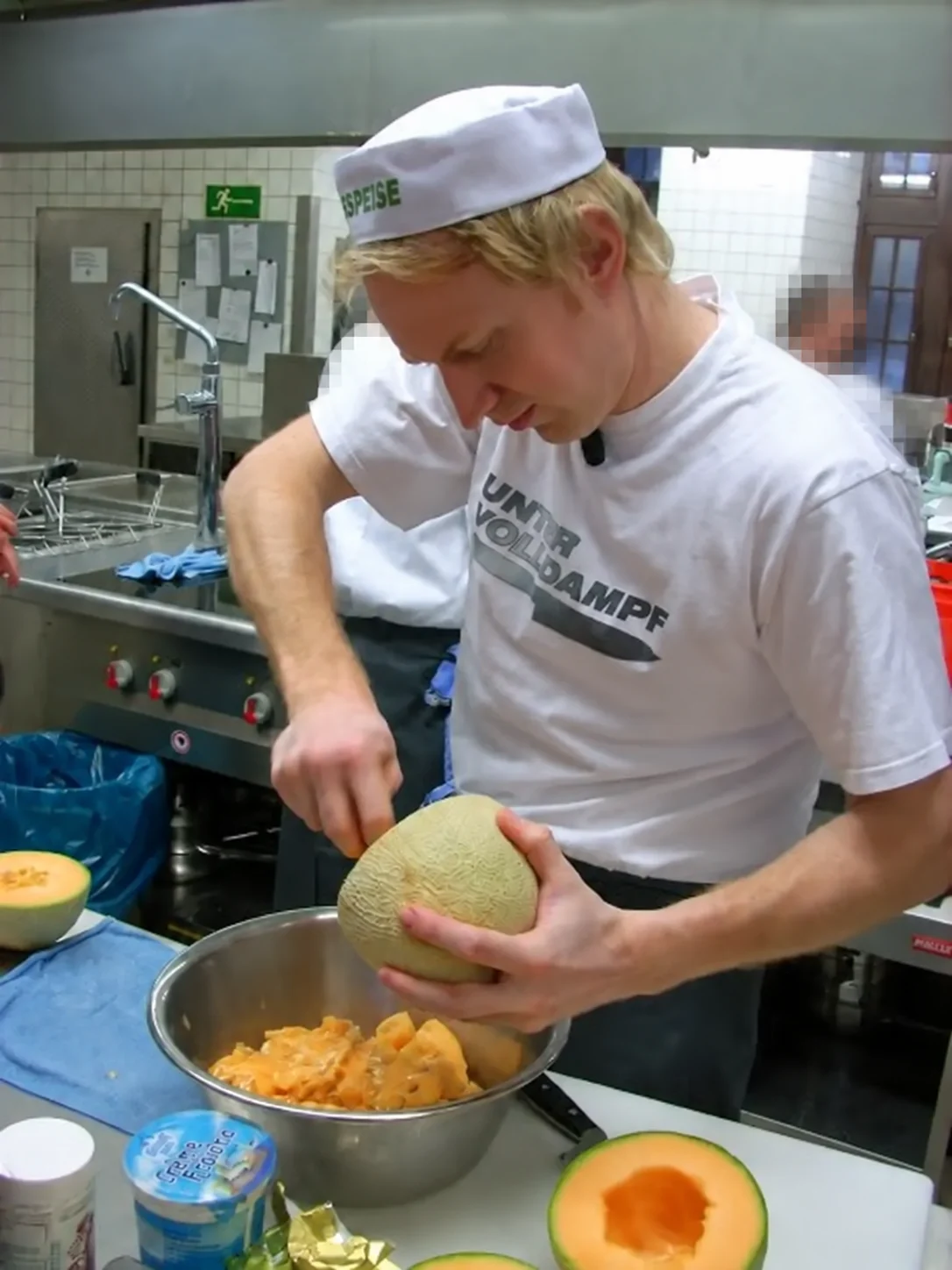 Marcus bereitet in der Küche eine Melone zu; eine weitere Person ist vollständig unscharf